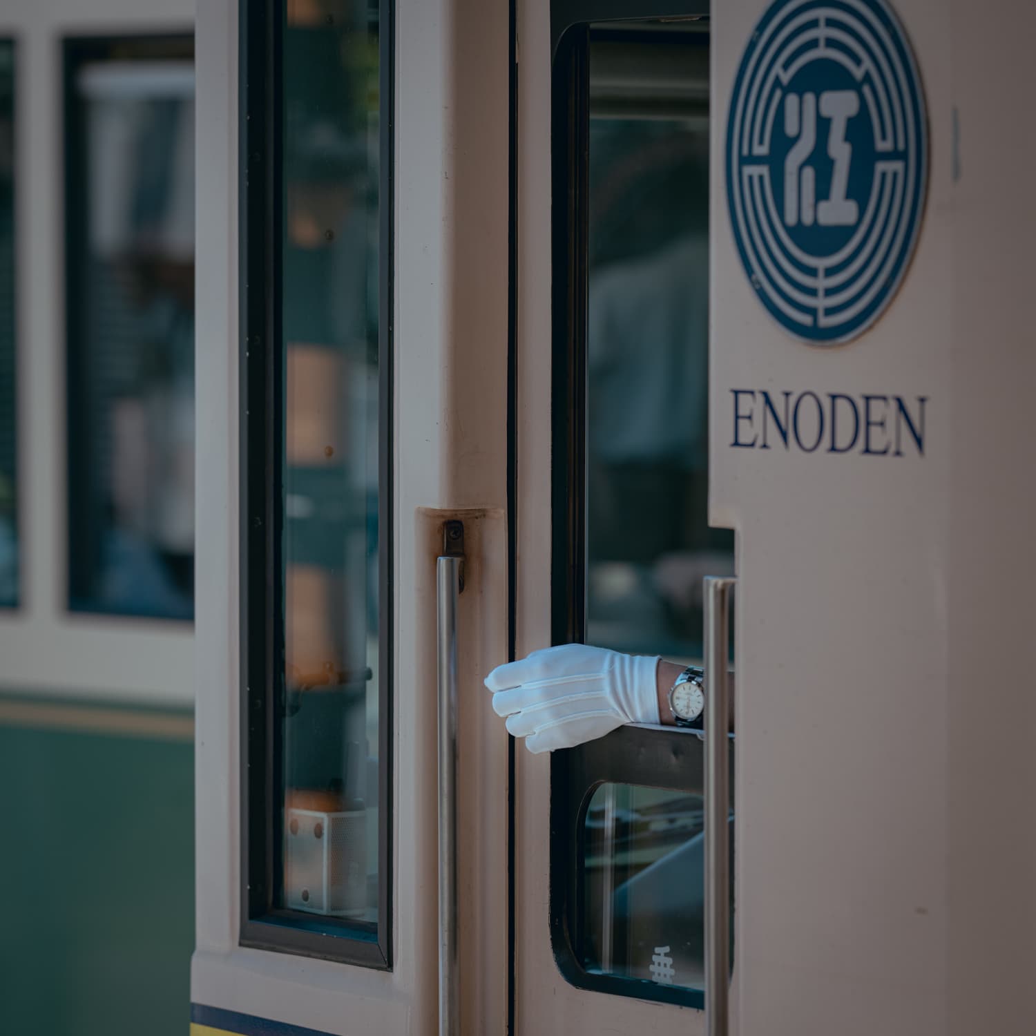 A close-up of a train door with a gloved hand extending out, featuring the ENODEN logo