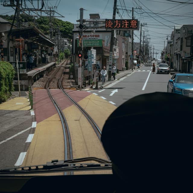 A view from inside a vehicle, looking out at a street with tram tracks running through it The tracks split into two directions, and there are buildings, power lines, and a sign with Japanese text in the background A person wearing a hat is partially visible in the foreground