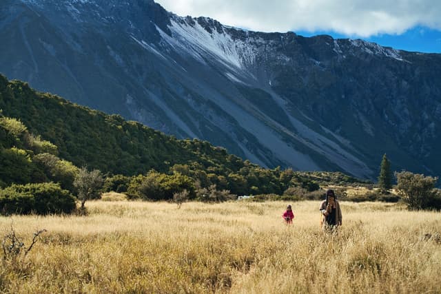 Two people walking through a grassy field with a backdrop of forested hills and a snow-capped mountain