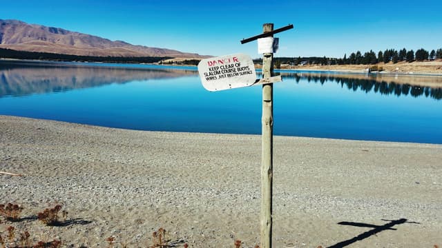 A serene lake with clear blue water, surrounded by a sandy shore and distant mountains A signpost in the foreground reads DANGER: NO SWIMMING