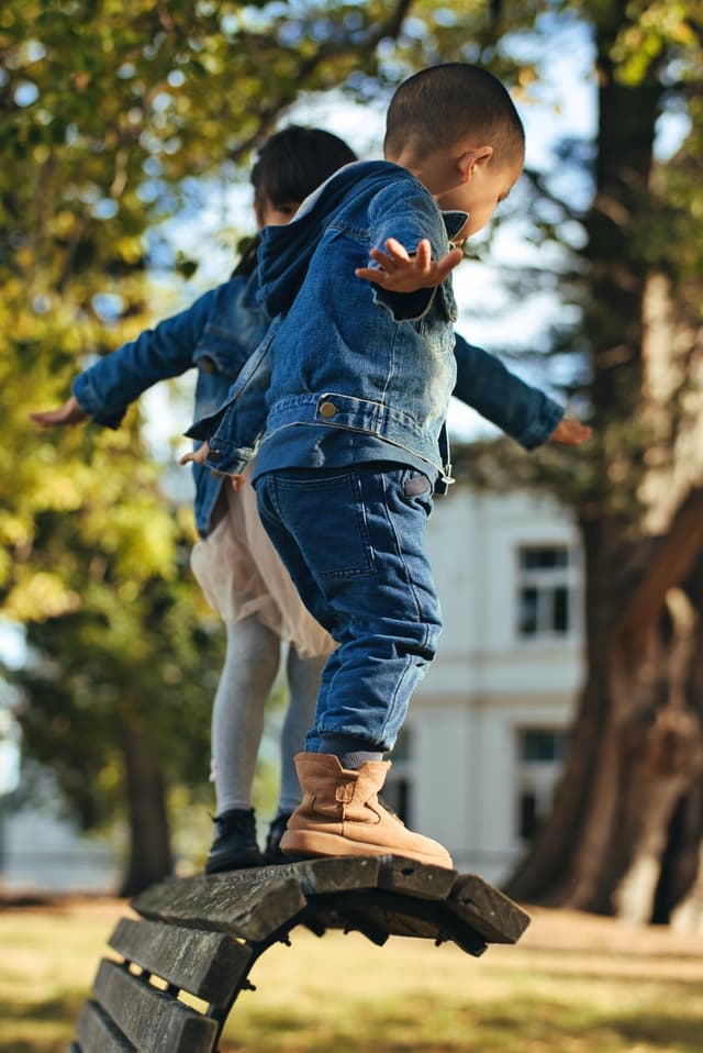 Two children balancing on the backrest of a park bench, arms outstretched, with trees and a building in the background