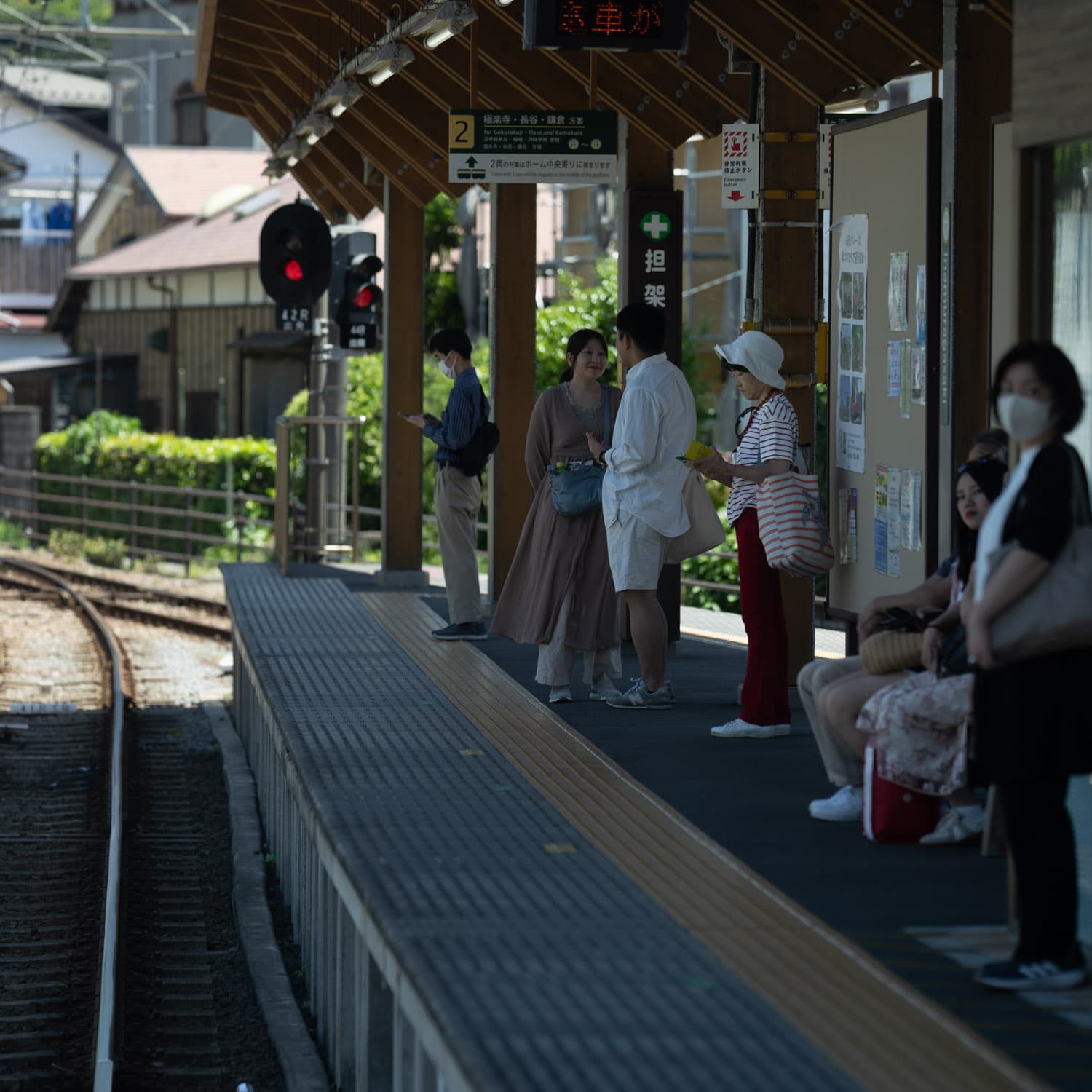 A train station platform with several people waiting, some sitting and some standing, under a wooden canopy The tracks curve to the left, and there are signal lights and a signboard in the background