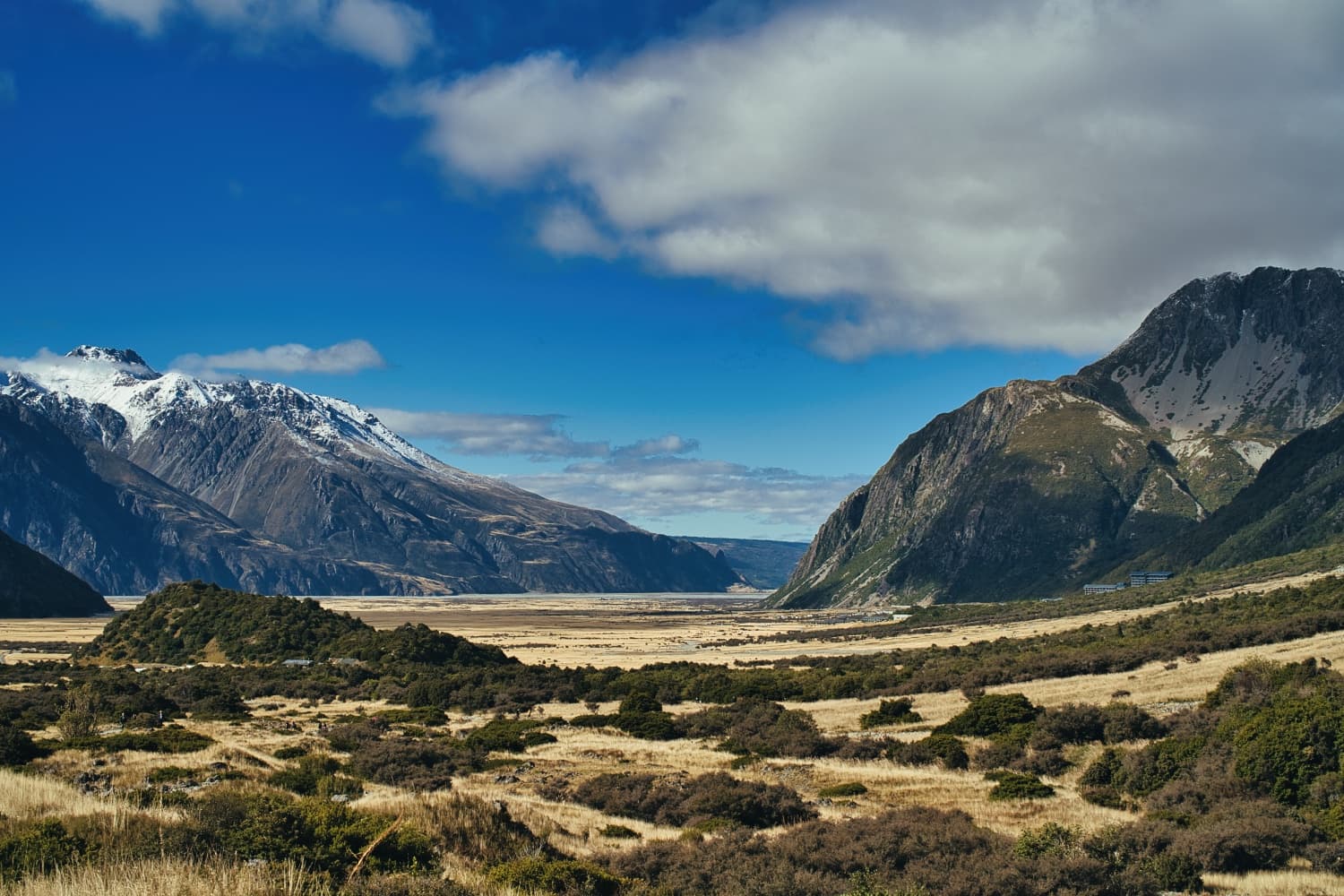 A scenic valley with snow-capped mountains in the background, grassy plains in the foreground, and a partly cloudy sky overhead