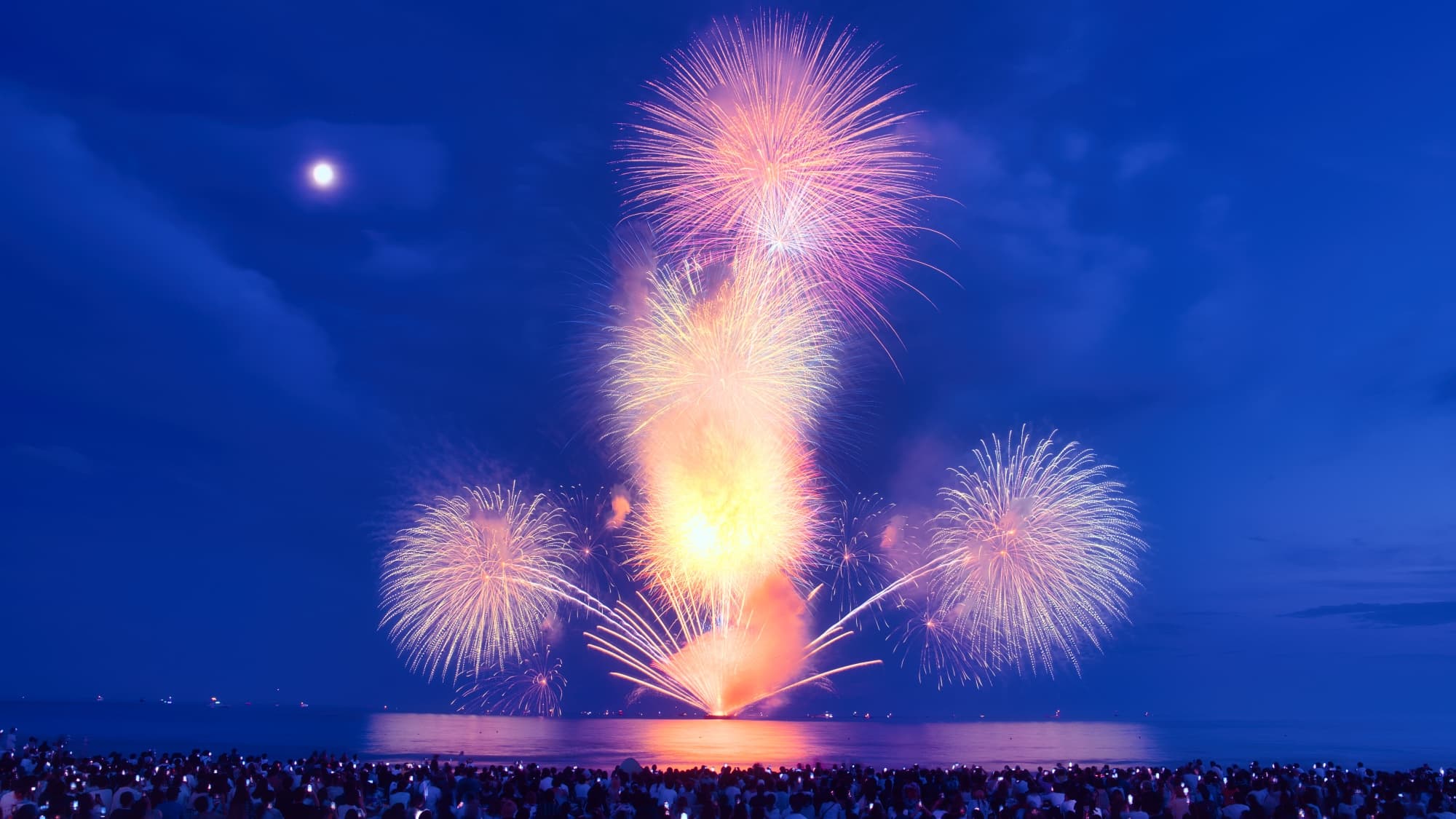 A vibrant fireworks display illuminates the night sky over a large body of water, with a crowd of spectators gathered along the shoreline