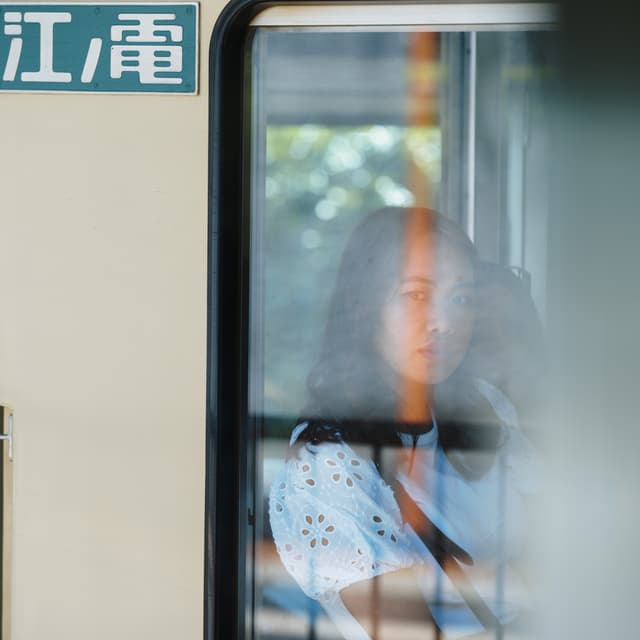 A person with long hair is seen through the window of a train, with a contemplative expression The background outside the train is blurred, and there is a sign with characters in a non-Latin script