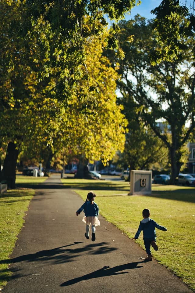 Two children running down a paved path in a park, surrounded by lush green grass and large trees with sunlight filtering through the leaves