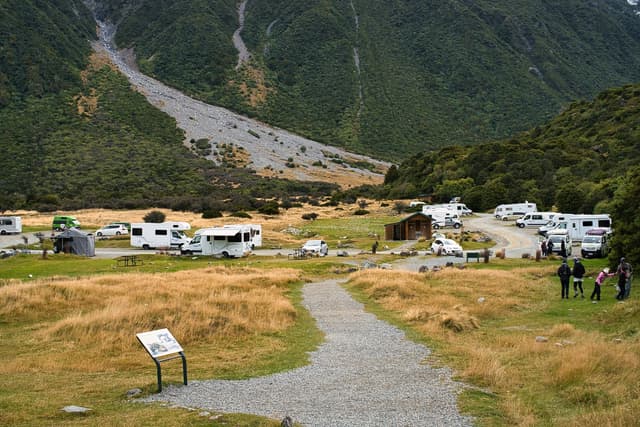 A gravel path leads through a grassy area towards a parking lot filled with camper vans, set against a backdrop of green hills and a rocky slope