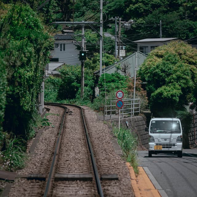 A railway track runs parallel to a narrow road with a small white vehicle driving on it, surrounded by lush greenery and residential houses in the background