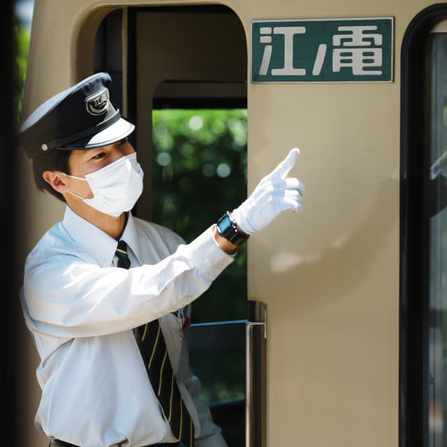 A uniformed train conductor wearing a face mask and white gloves is standing at the door of a train, pointing in a direction The train has a sign with Japanese characters