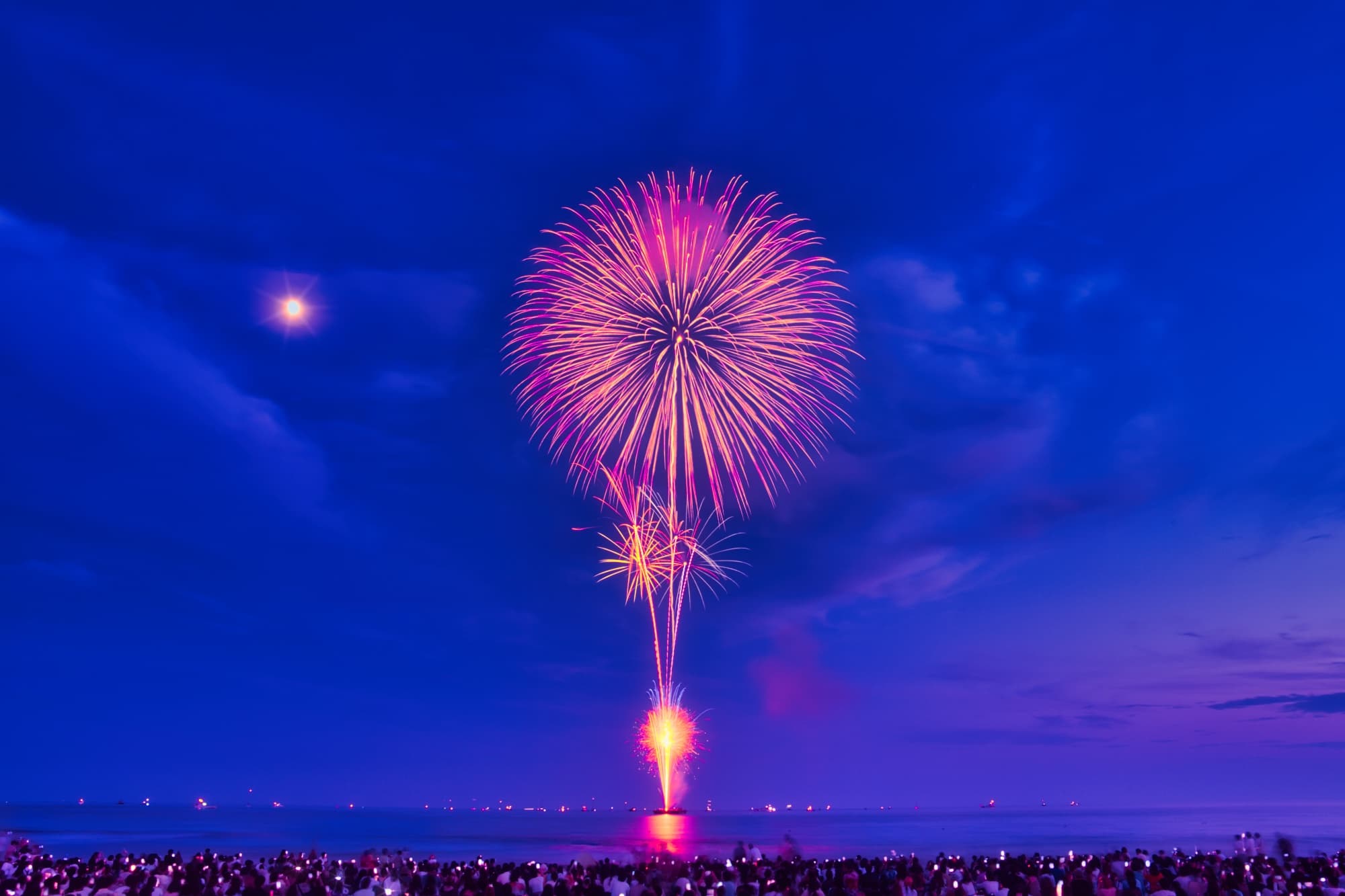 A vibrant firework display illuminates the night sky over a large crowd, with a bright moon visible in the background