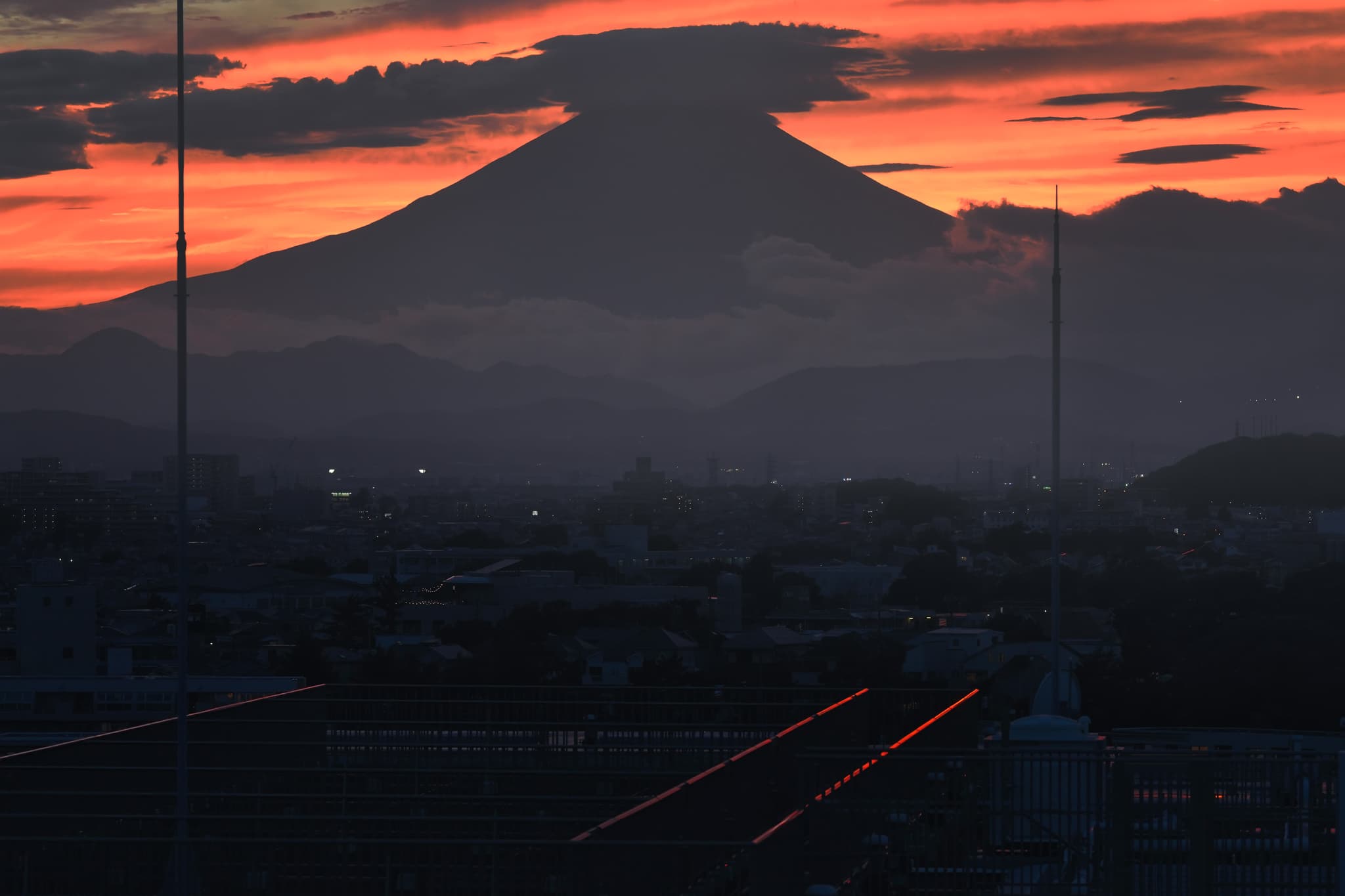 A silhouetted mountain against a vibrant orange and red sunset sky, with a cityscape in the foreground and faint lights visible