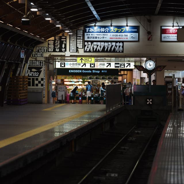 A dimly lit train station platform with tracks in the foreground, a convenience store, and various signs in Japanese A clock is visible on the wall, and a few people are gathered near the store