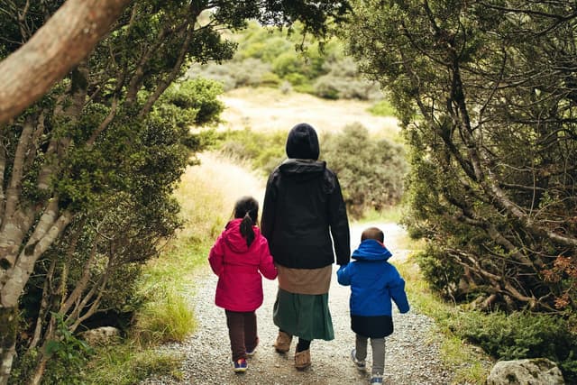 An adult and two children walking on a gravel path surrounded by trees, with the children wearing colorful jackets