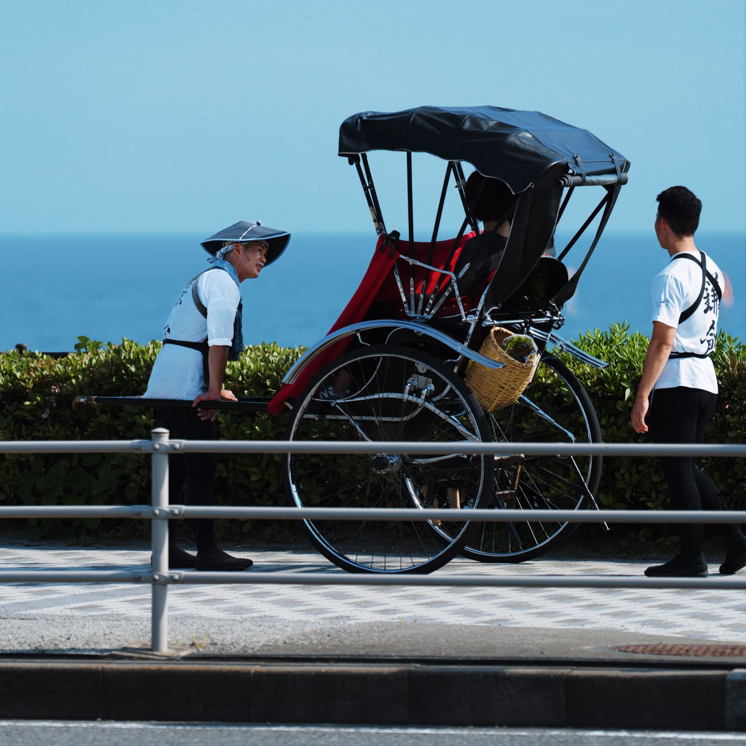 A rickshaw being pulled by a person in traditional attire, with another person standing nearby, set against a backdrop of the ocean and a clear blue sky